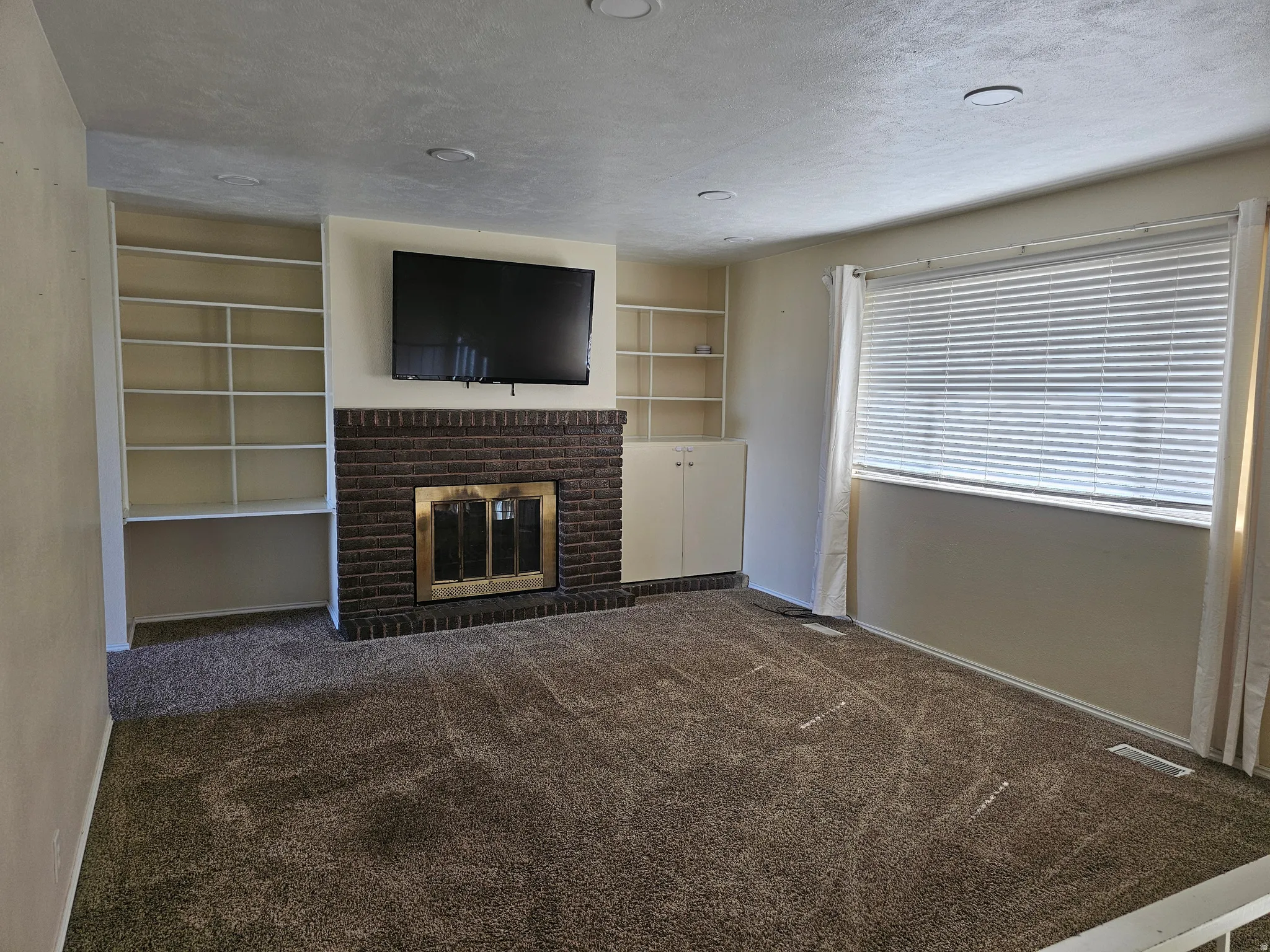 Brick fireplace with brass insert, built-in shelving, carpet flooring, recessed lighting, and window with blinds