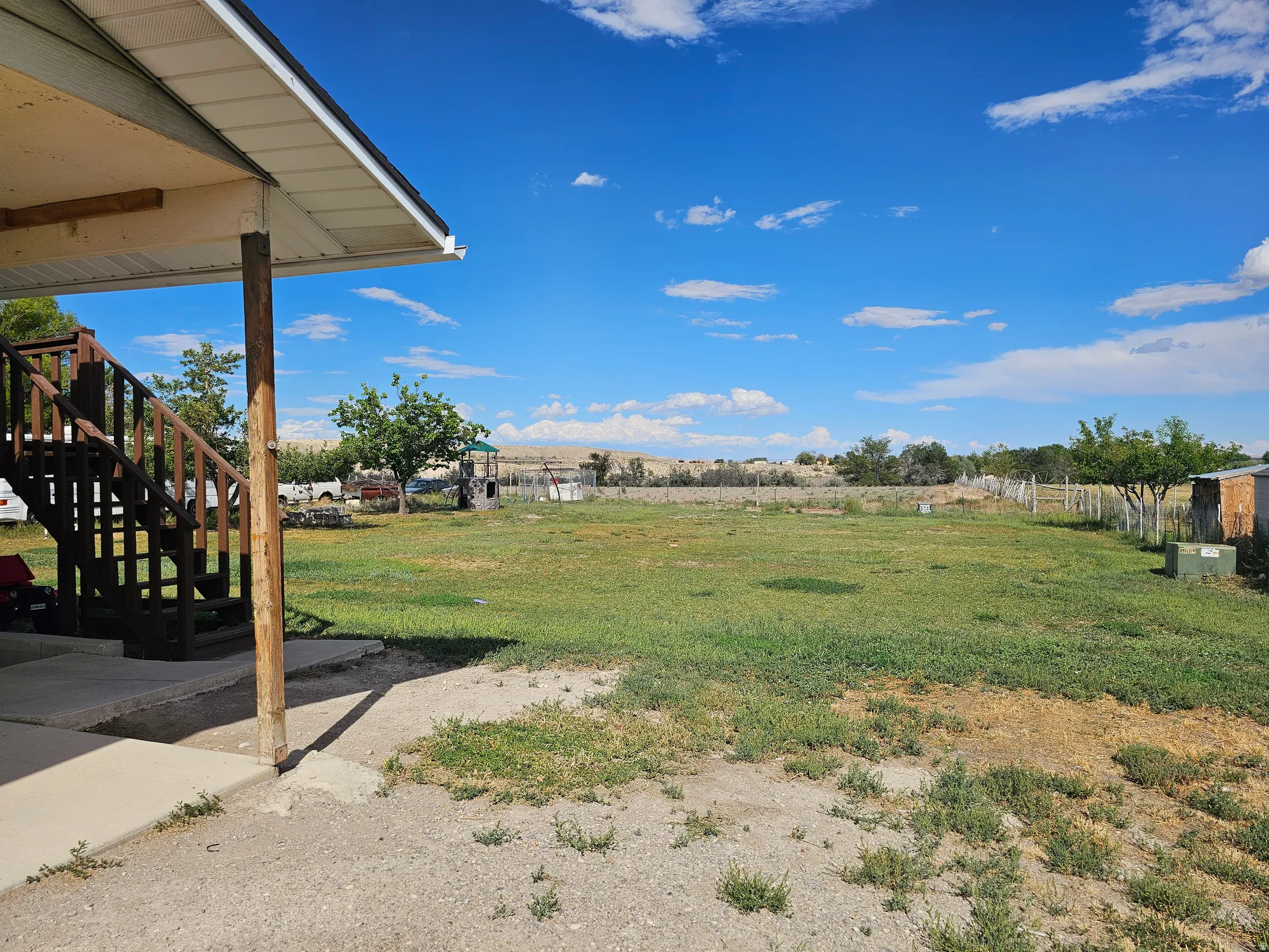 Expansive rural property featuring open acreage with mature landscaping, covered porch with wooden support posts, and distant countryside backdrop