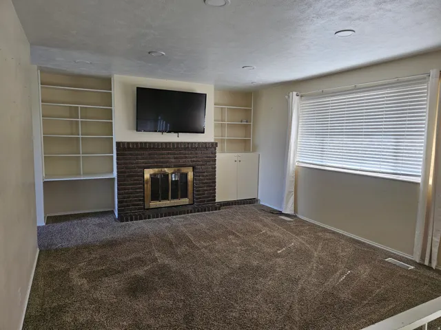 Brick fireplace with brass insert, built-in shelving, carpet flooring, recessed lighting, and window with blinds