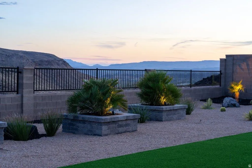Expansive outdoor entertaining space featuring manicured lawn, illuminated specimen palms in planter beds, decorative hardscape with gravel finish, and metal railing along the perimeter