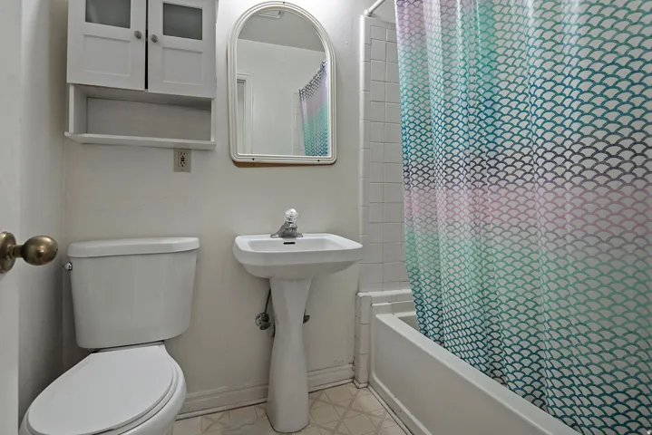 Pedestal sink with chrome faucet, white toilet, built-in white wall cabinet, white bathtub with tiled surround, and patterned flooring