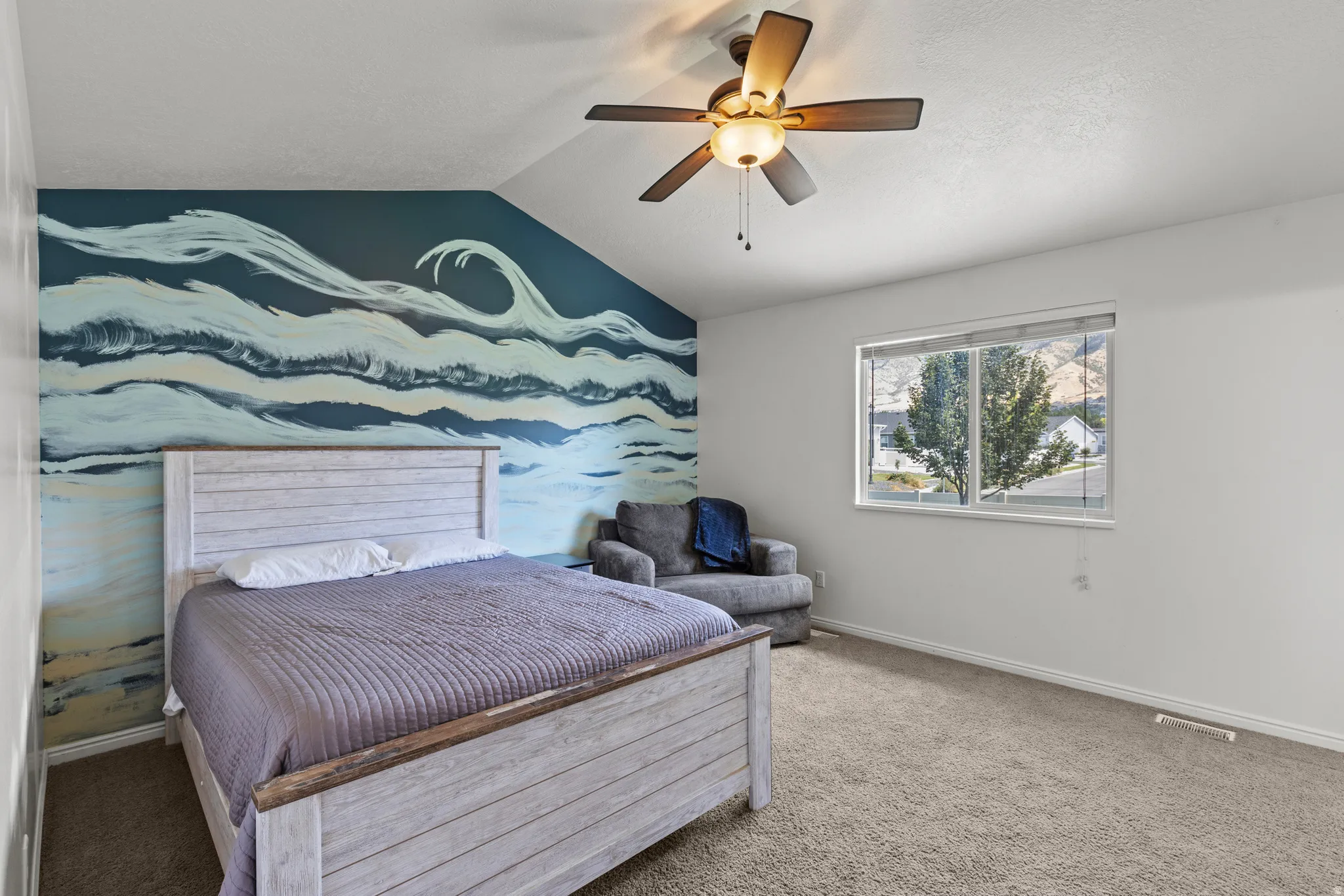 Spacious primary bedroom featuring a custom wave-pattern accent wall in cool tones, wood-finish platform bed frame, ceiling fan with integrated lighting, and carpet flooring