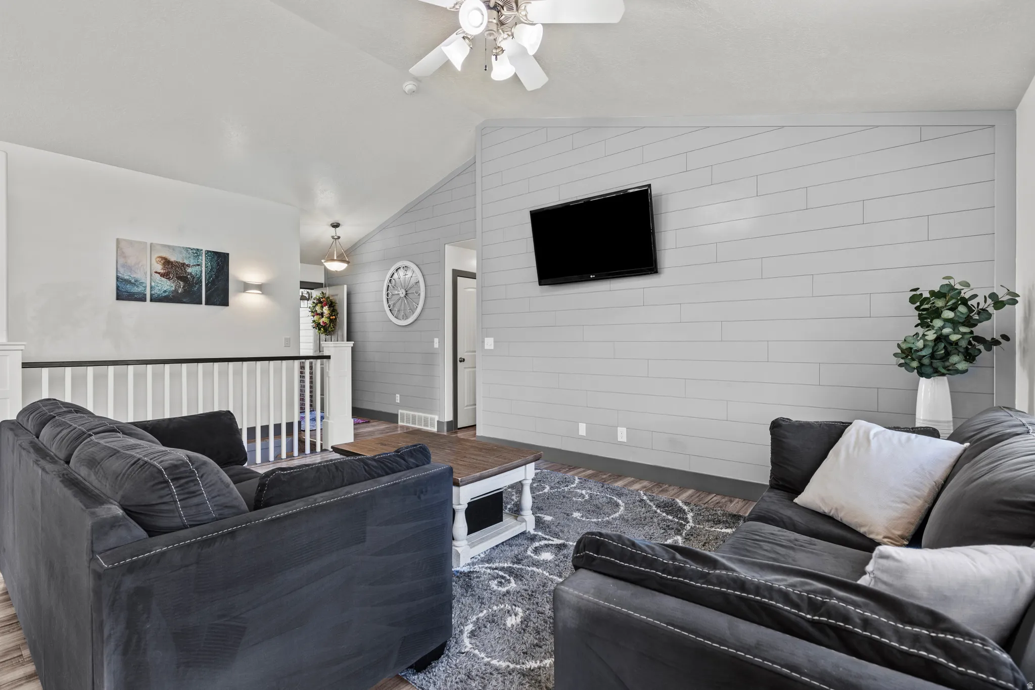 Vaulted ceiling, shiplap accent wall, wood-finish flooring, white spindle railing with a dark top rail, and a ceiling fan with integrated lighting