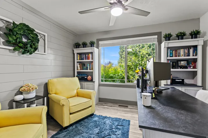 Bright, light-filled home office featuring built-in shelving on either side of a glass door, hardwood flooring, a ceiling fan with integrated lighting, white shiplap accent wall, and a dark countertop workspace with integrated storage