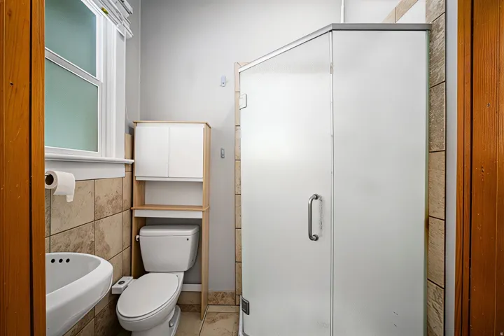 Walk-in shower with frosted glass enclosure, tile surround, wall-mounted sink, and a window with blinds