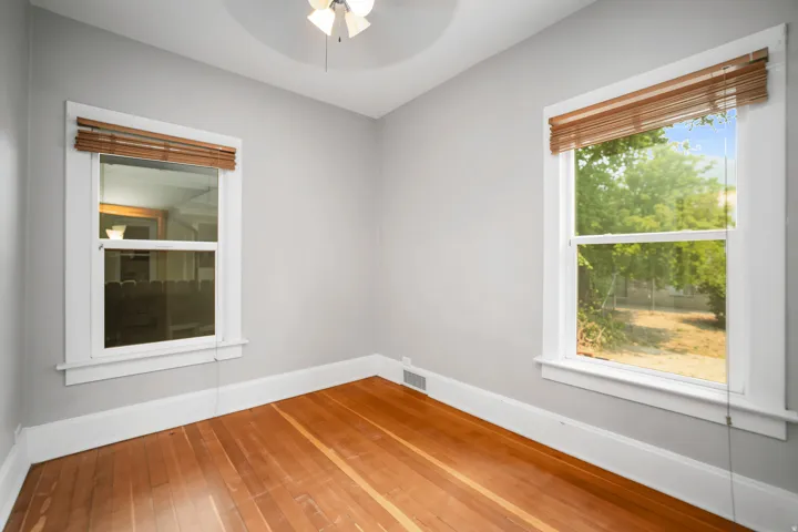 Hardwood flooring, two windows with white trim, a ceiling fan, and light gray walls