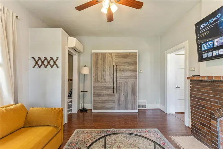 Wood-finish flooring with a brick fireplace, ceiling fan, and wall-mounted air conditioning unit