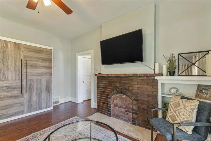 Exposed brick fireplace with an arched hearth, built-in shelving, wood-finish flooring, ceiling fan, and paneled storage unit