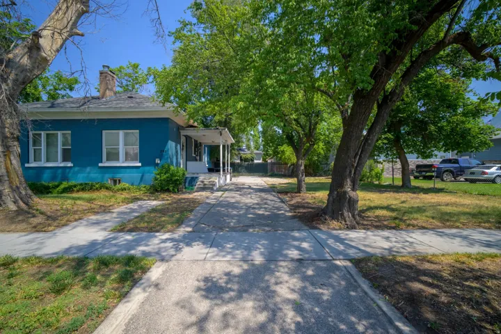 Blue exterior with white trim, covered front porch, brick chimney, paved driveway, and mature trees