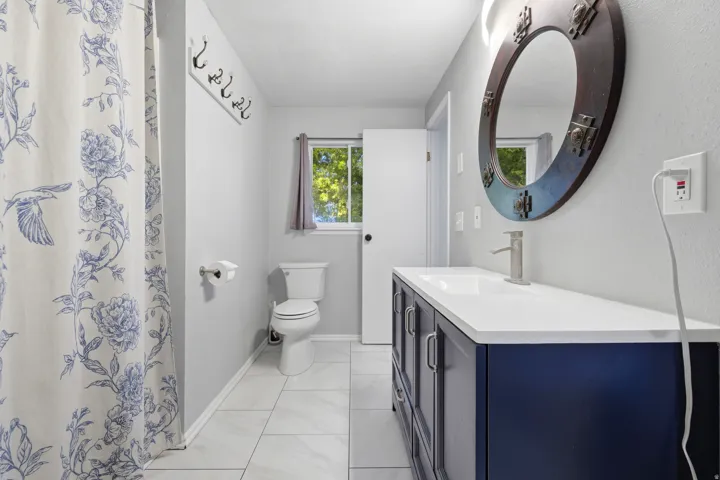White tile flooring, a dark blue vanity with a white countertop, a circular framed mirror, and a window with a curtain