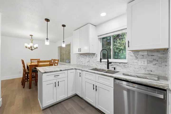 White shaker cabinetry, light-toned countertops, tile backsplash, stainless steel dishwasher, and wood-finish flooring