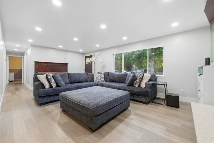 Spacious living room anchored by a sectional seating arrangement and upholstered ottoman, featuring recessed ceiling lighting, wood-finish flooring throughout, and expansive windows framing natural landscape views