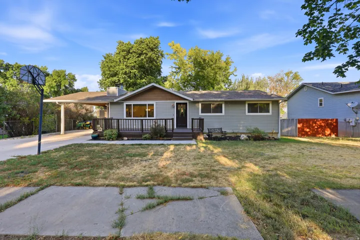 Exterior featuring light gray siding, dark wood-finish deck, covered carport, front yard, and a paved driveway
