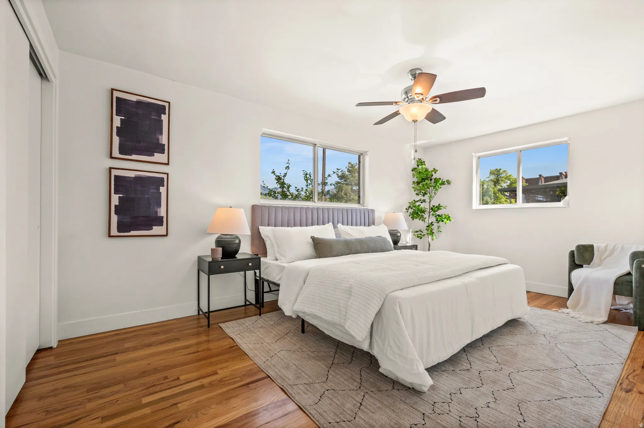 Bright primary bedroom showcasing hardwood flooring, ceiling fan with integrated lighting, and abundant natural light from multiple windows