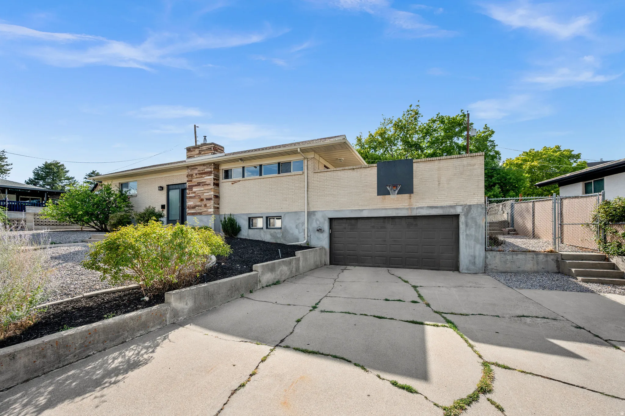 Modern architectural home featuring a sleek concrete driveway with linear design elements, dark-finish garage door, mixed-material exterior combining stucco and wood-finish accents, and mature landscaping with native plantings and established trees throug