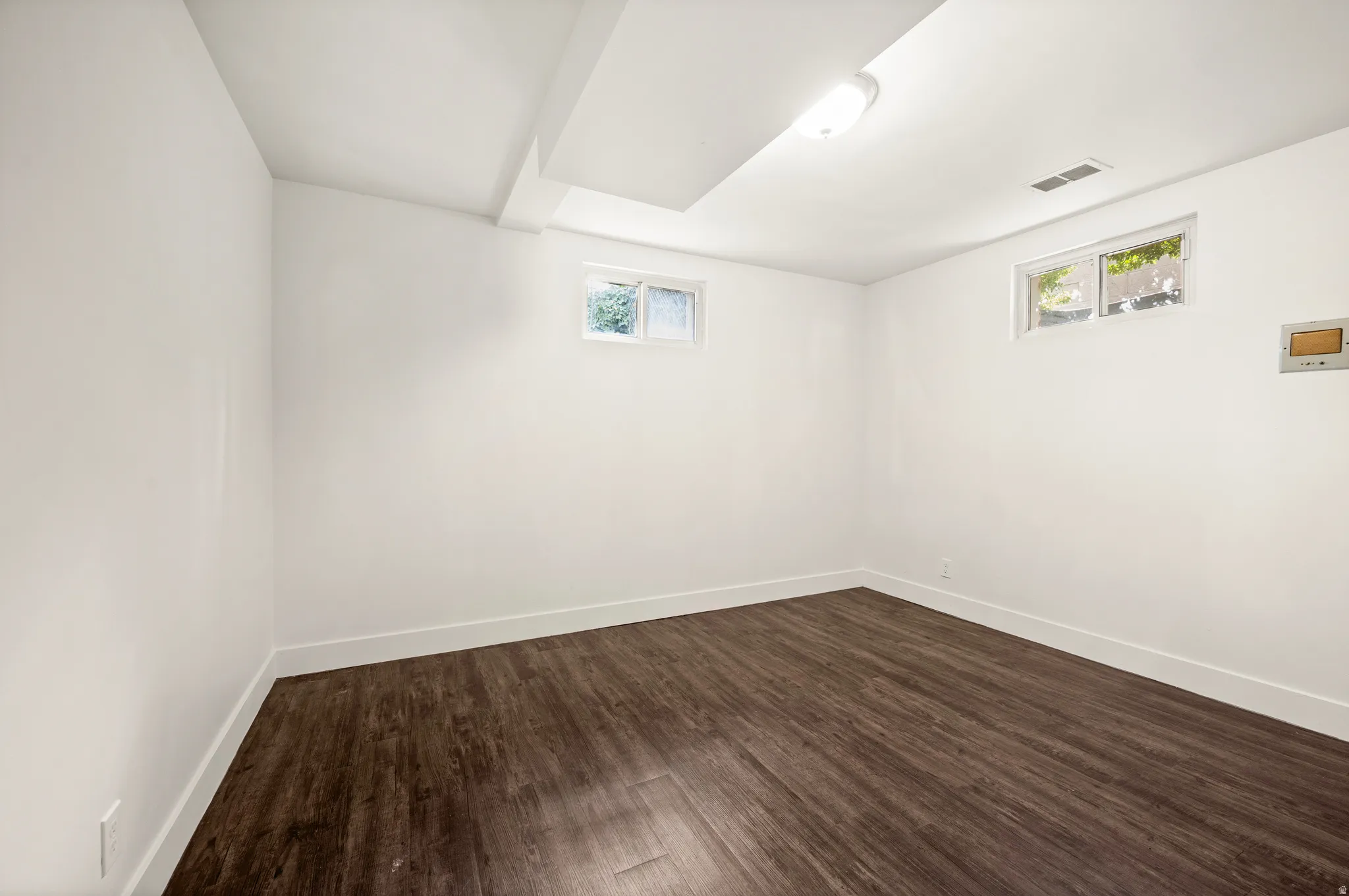 Bright, generously proportioned room featuring dark wood-finish flooring, clean white walls, and recessed ceiling lighting
