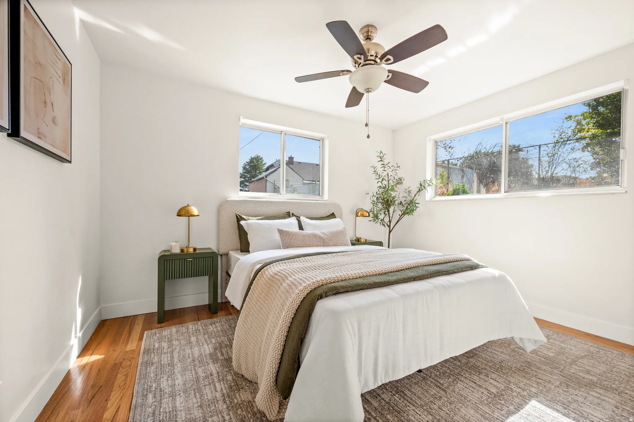 Bright bedroom showcasing wood-finish flooring, abundant natural light from multiple windows, ceiling fan with integrated lighting, and neutral wall finishes