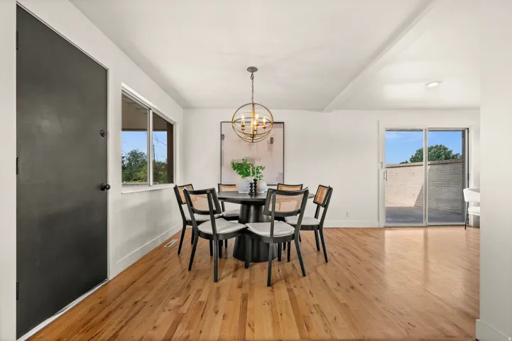 Bright dining area showcasing gleaming wood-finish flooring, abundant natural light from multiple windows, glass sliding doors opening to an outdoor patio, and a modern pendant fixture overhead