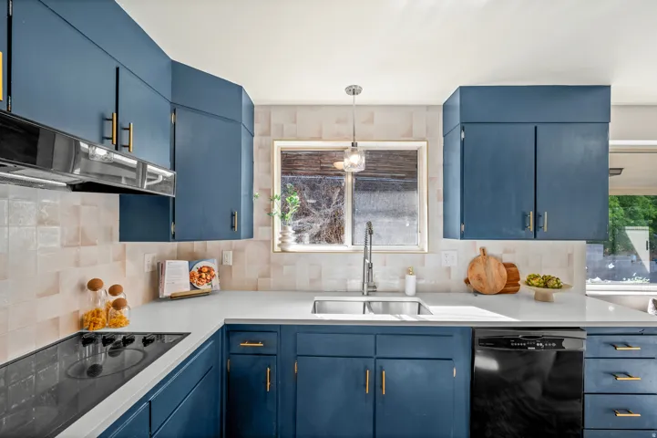 Modern kitchen featuring custom cabinetry in deep blue with brass hardware, stainless steel appliances including a black dishwasher, quartz countertops, a ceramic tile backsplash, and a stainless steel undermount sink with contemporary faucet