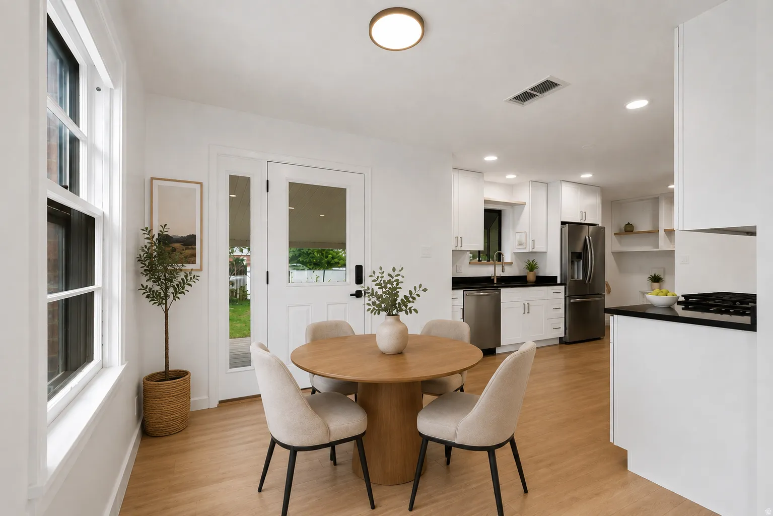 Open-concept kitchen and dining area featuring warm wood-finish flooring, a round wood dining table with seating for four, white cabinetry with black countertops and stainless steel appliances, abundant natural light from multi-pane windows and glass door