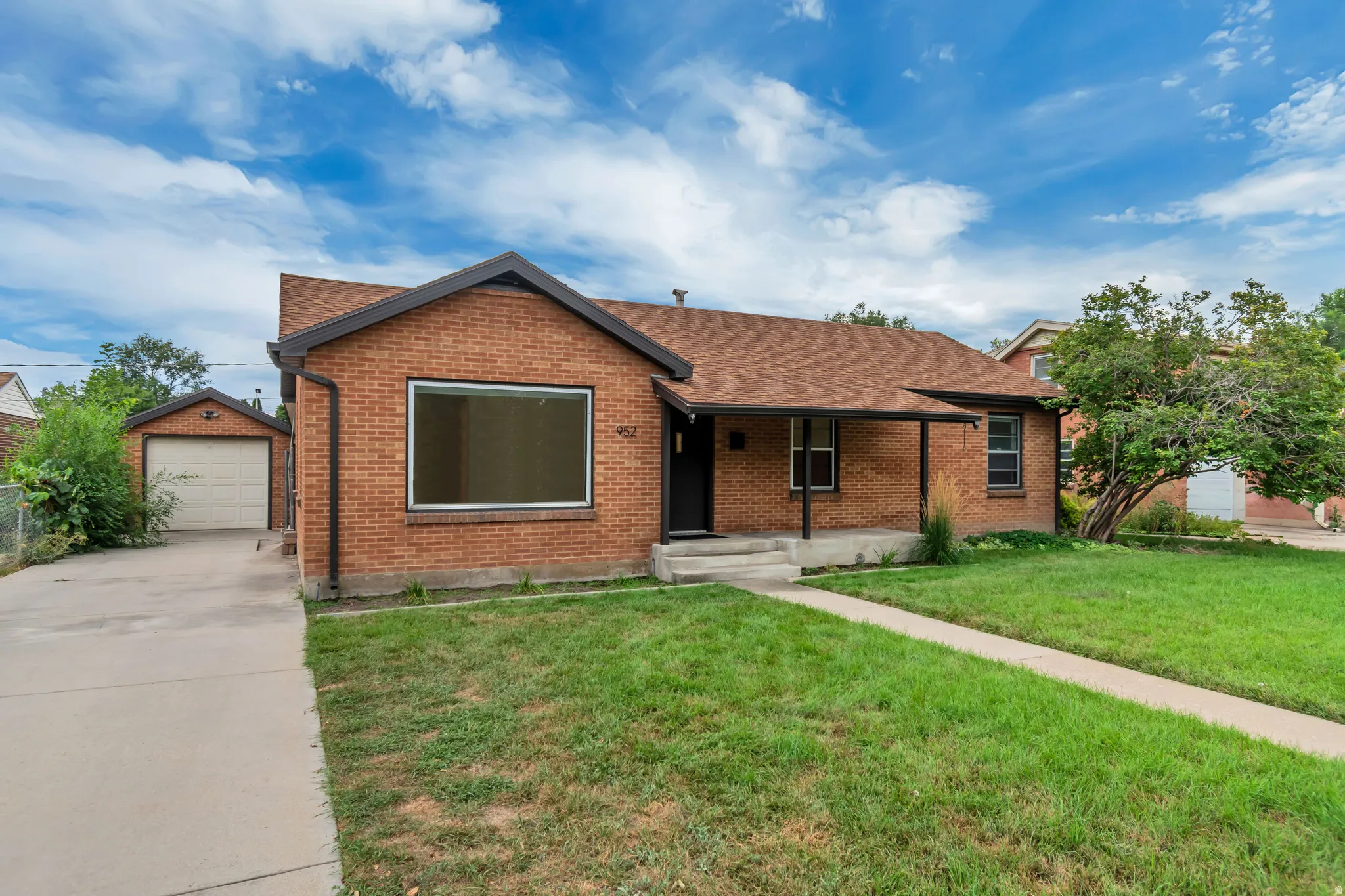 Brick Ranch home featuring a large picture window, attached single-car garage, concrete driveway, and mature landscaping with established trees and manicured lawn