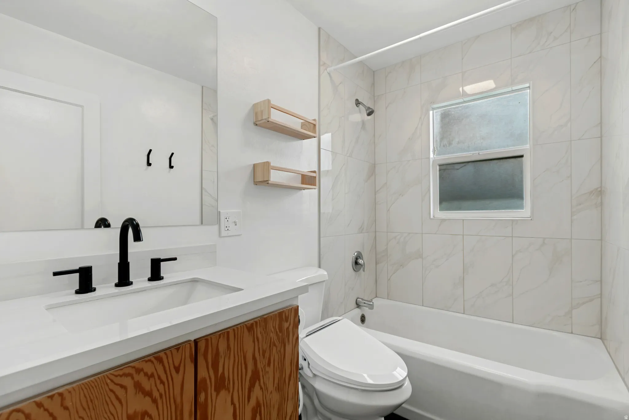 Modern bathroom featuring a wood-finish vanity with integrated sink, white quartz countertop, matte black faucet and hardware, large framed mirror, marble-look tile surround, walk-in shower with glass enclosure, soaking tub, and natural light from operabl