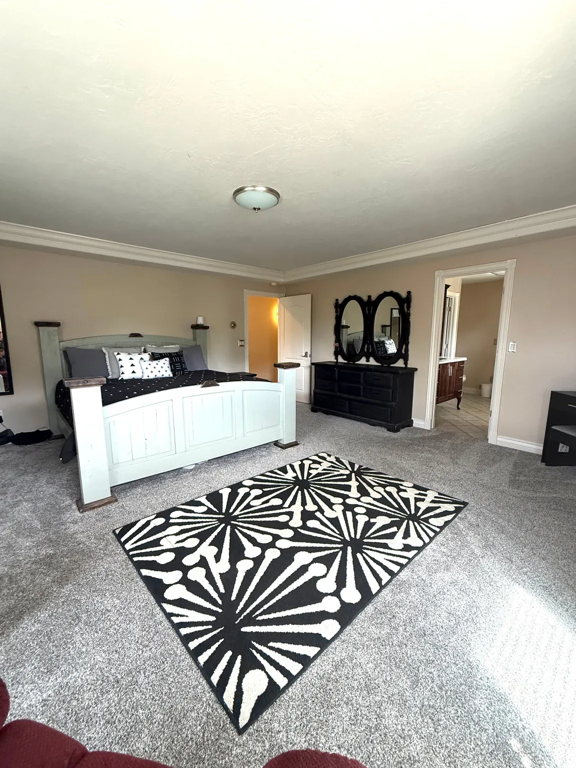 Spacious primary bedroom suite featuring a white-finished bed frame, gray carpeting throughout, recessed ceiling lighting, and a geometric patterned area rug