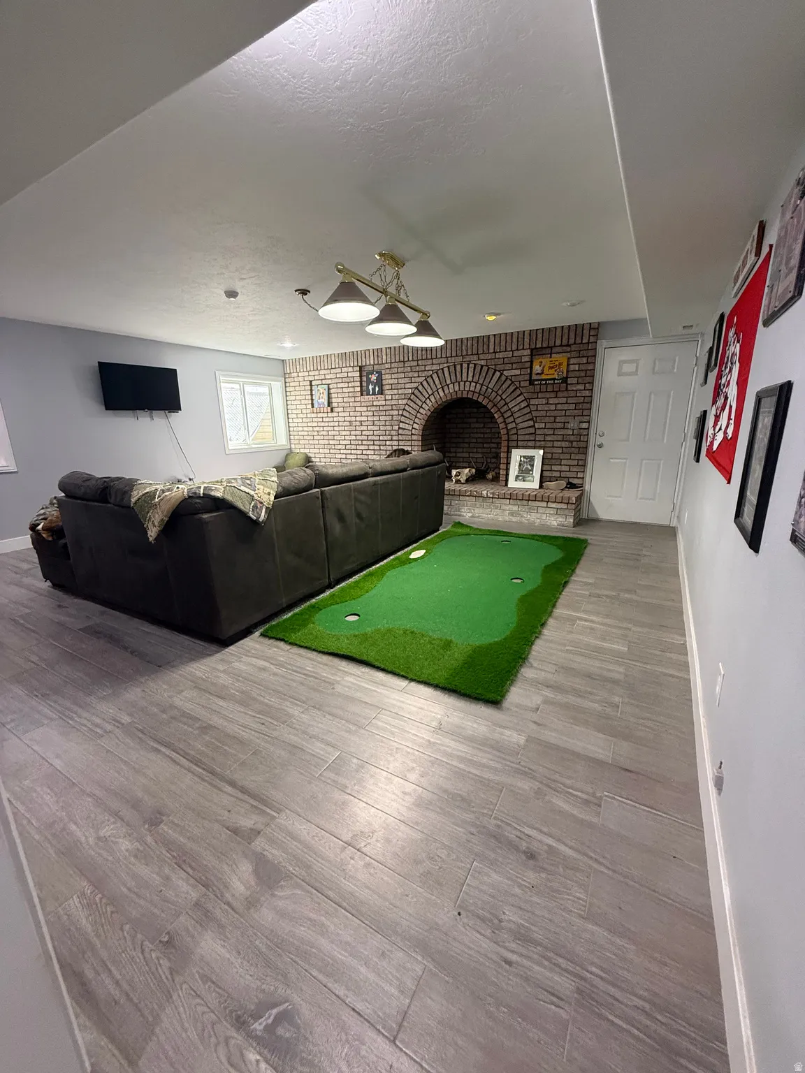 Finished basement recreation space featuring wood-finish plank flooring, exposed brick fireplace with arched opening, vaulted ceiling with recessed lighting, built-in seating area, and dedicated putting green with synthetic turf surface