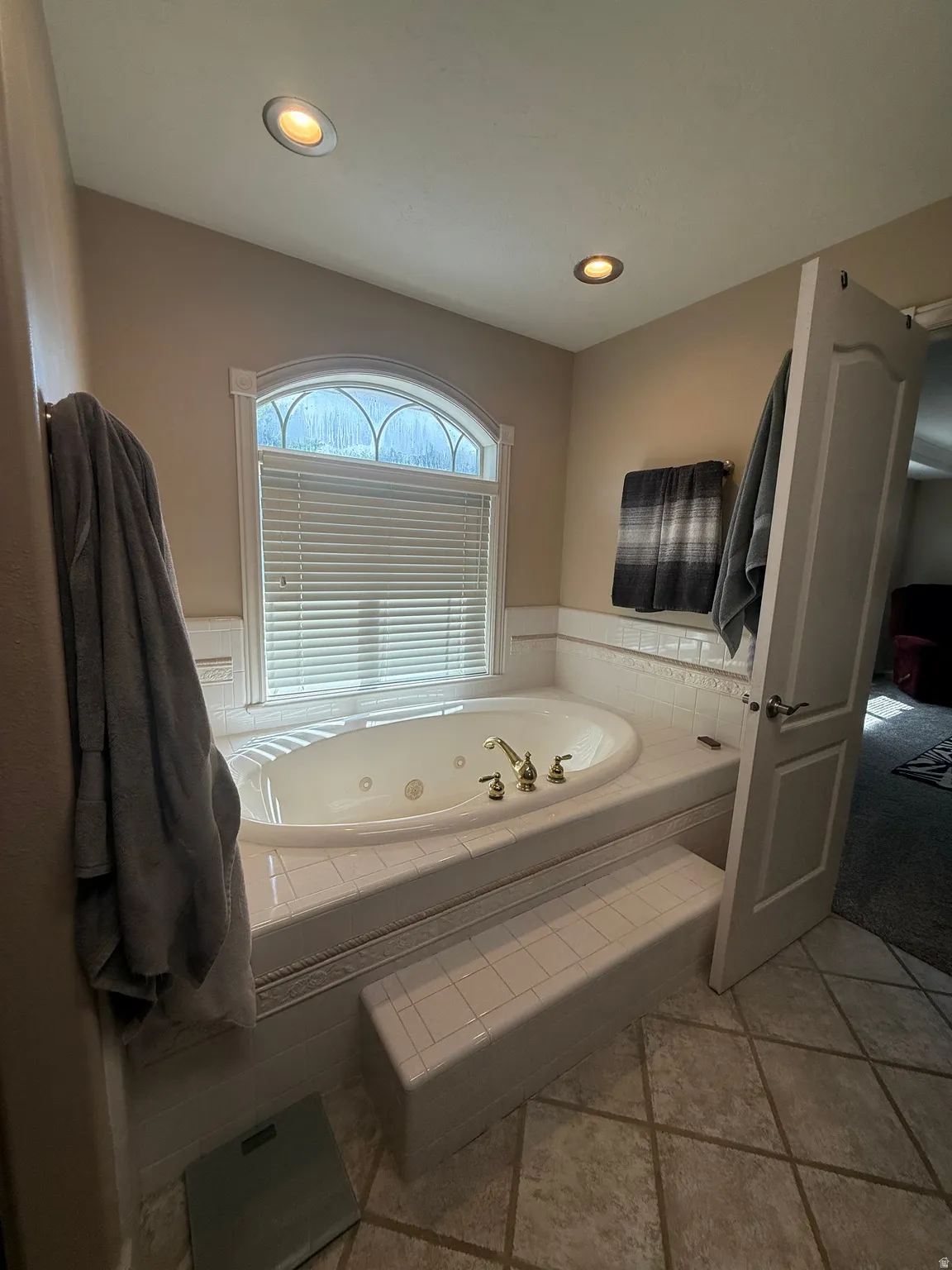 Recessed lighting illuminates the jetted tub with brass fixtures, set within a tiled surround and an arched window