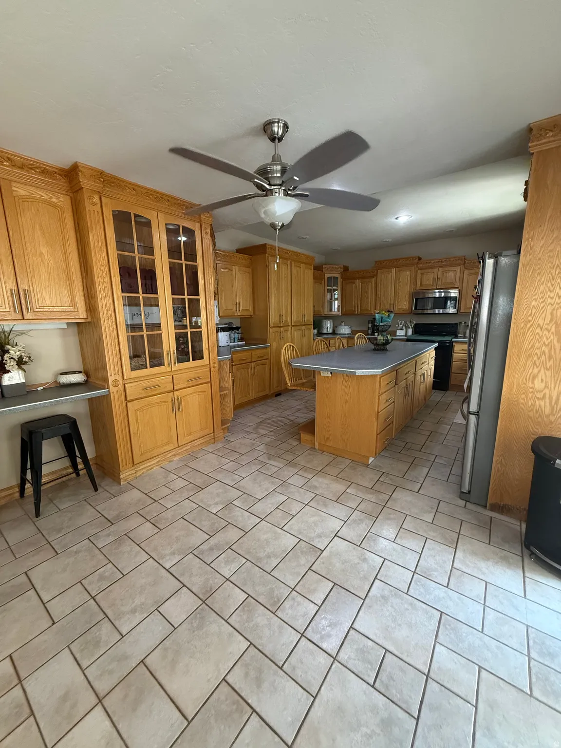 Kitchen featuring extensive light wood cabinetry, a central island with a countertop, integrated stainless steel appliances, a ceiling fan with integrated lighting, and tile flooring