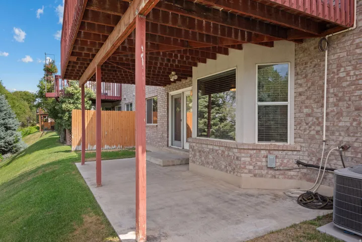 Covered concrete patio with a wood-framed deck overhead, brick exterior, and multiple windows