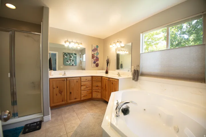 Spa-inspired master bath featuring dual vanities with wood-finish cabinetry, paired mirrors with integrated lighting, tile flooring, a soaking tub with deck-mounted fixtures, and a walk-in shower enclosure with glass doors