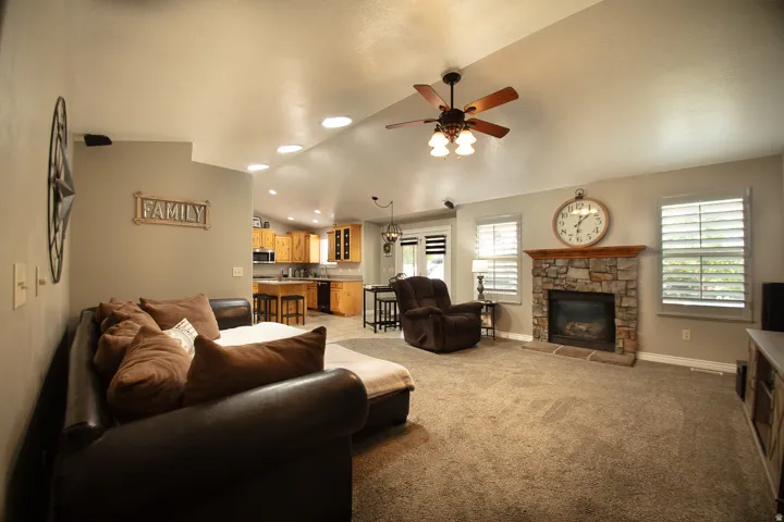 Vaulted ceiling with recessed lighting, stone-faced fireplace with wood mantel, wood-finish kitchen cabinetry, and ceiling fan with integrated lighting