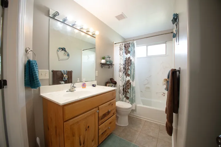Wood-finish vanity with a white countertop and integrated sink, large vanity mirror with overhead lighting, white toilet, and a white bathtub with a window above