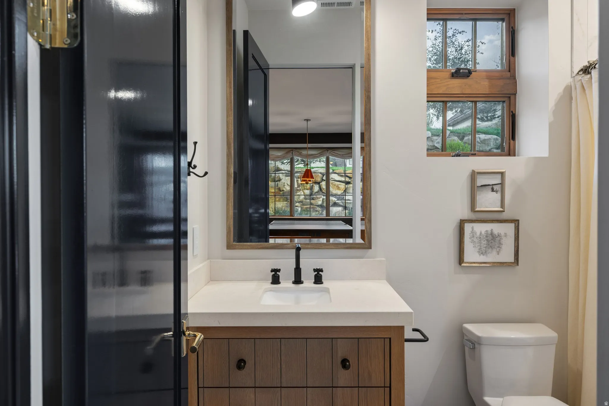 Contemporary bathroom featuring a wood-finish vanity with dual-handle matte black faucet and white countertop, framed mirror with integrated lighting, black-framed glass shower enclosure, and white porcelain toilet