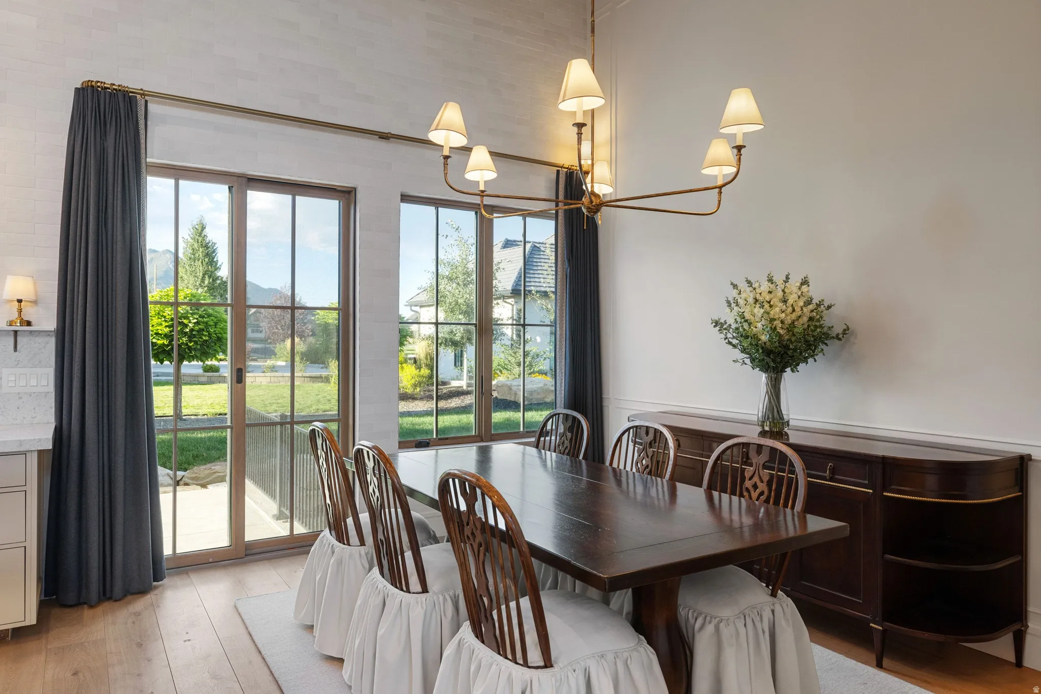 Elegant dining room featuring French doors with multiple panes opening to manicured grounds, wood-finish flooring, a dark wood dining table, coordinating Windsor-style chairs with slipcovers, a brass multi-arm chandelier with fabric shades, wall sconces,