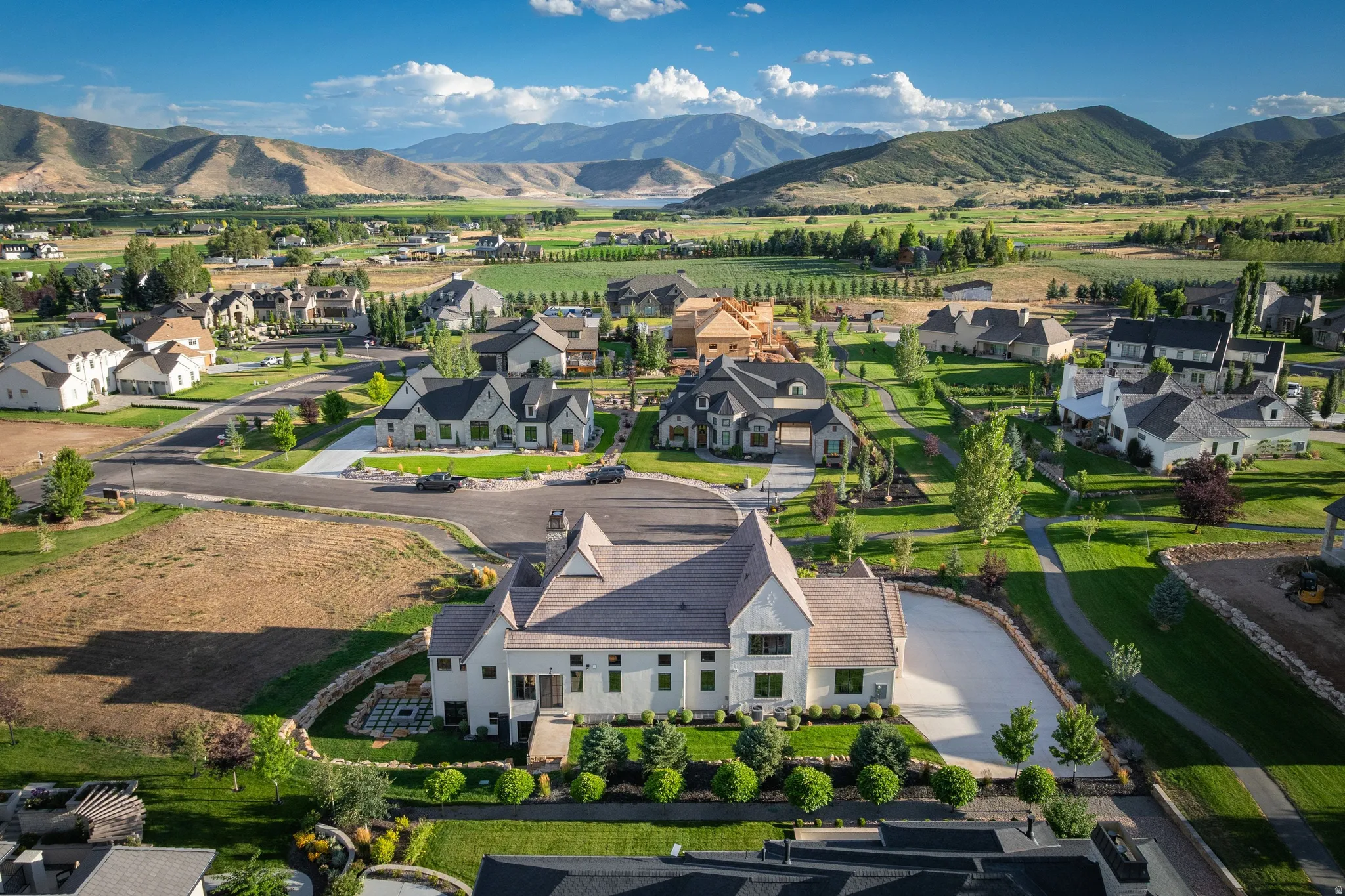 Elegant residential estate set on expansive grounds featuring a distinctive peaked-roof architectural design with light-colored exterior, mature landscaping with manicured lawn and established trees, curved driveway with paved entry, and mountain vistas a