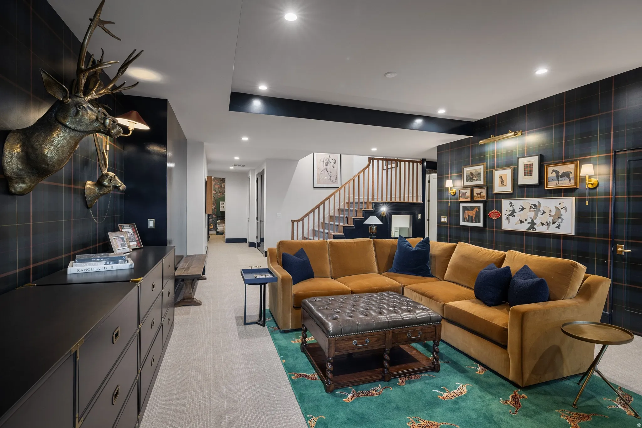 Sophisticated recreation room showcasing a curved mustard-toned sectional, dark plaid accent wall with integrated shelving and display niches, wood-finish flooring, recessed ceiling lighting, and a leather upholstered ottoman coffee table