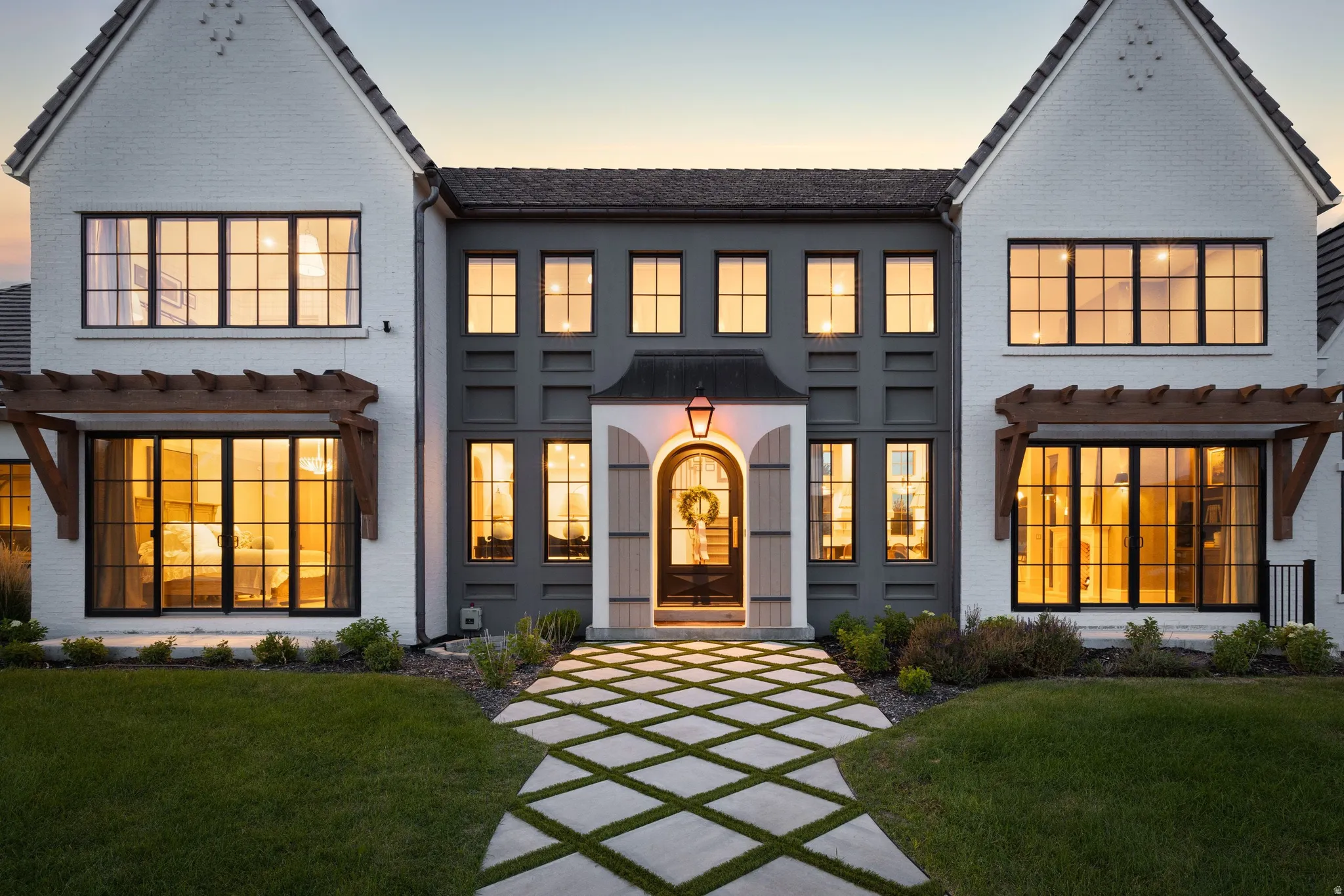 Modern Farmhouse estate showcasing a symmetrical facade with white and charcoal exterior finishes, multiple multi-pane windows with warm interior lighting, gabled rooflines with decorative trim details, a centered arched entryway with lantern sconce, and