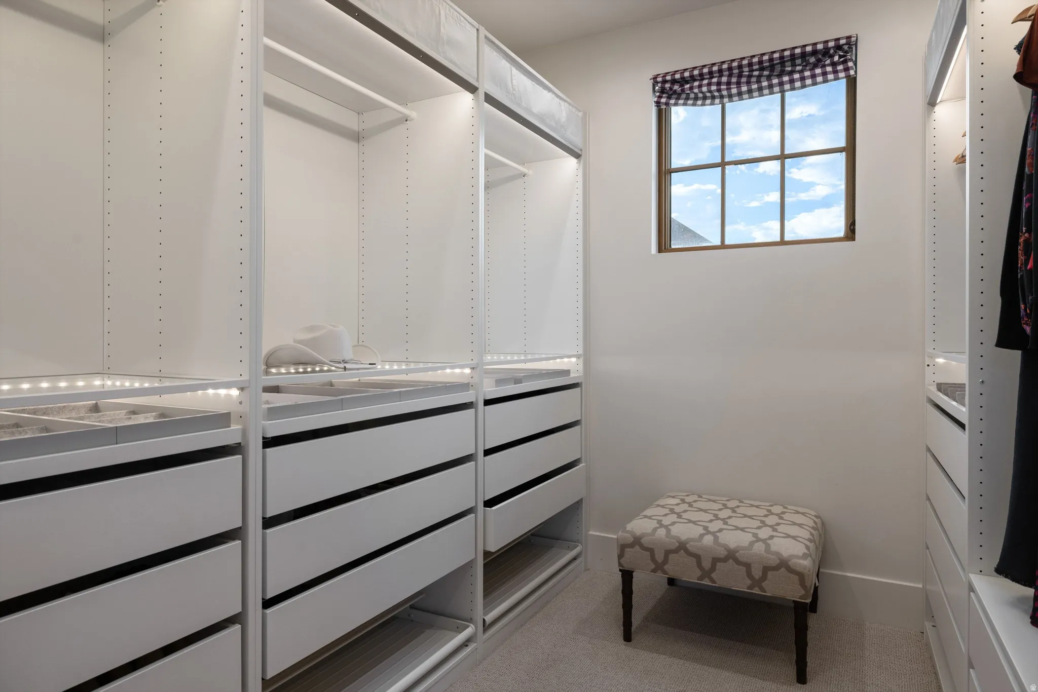 Luxurious walk-in closet featuring mirrored sliding doors, extensive shelving and drawer storage with contrasting hardware accents, and abundant natural light from a large window with checkered valance
