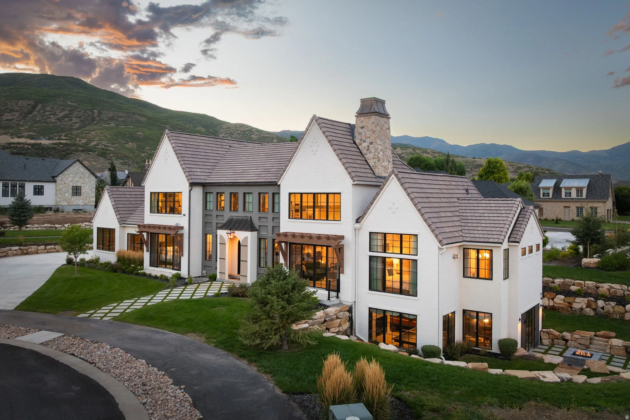 Modern farmhouse estate featuring a striking white exterior with multiple peaked rooflines and a tall stone chimney, expansive multi-pane windows with warm interior lighting, manicured lawn with ornamental grasses, and a circular driveway with permeable p