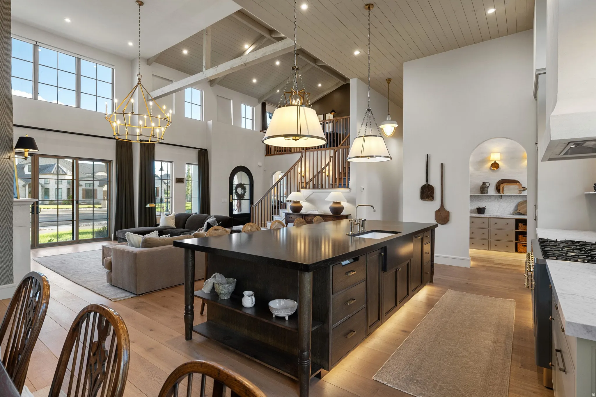 Open-concept kitchen and living area showcasing a black center island with storage and seating, stainless steel appliances, wood-finish flooring throughout, vaulted ceilings with exposed beams and recessed lighting, and a grand staircase with wrought iron