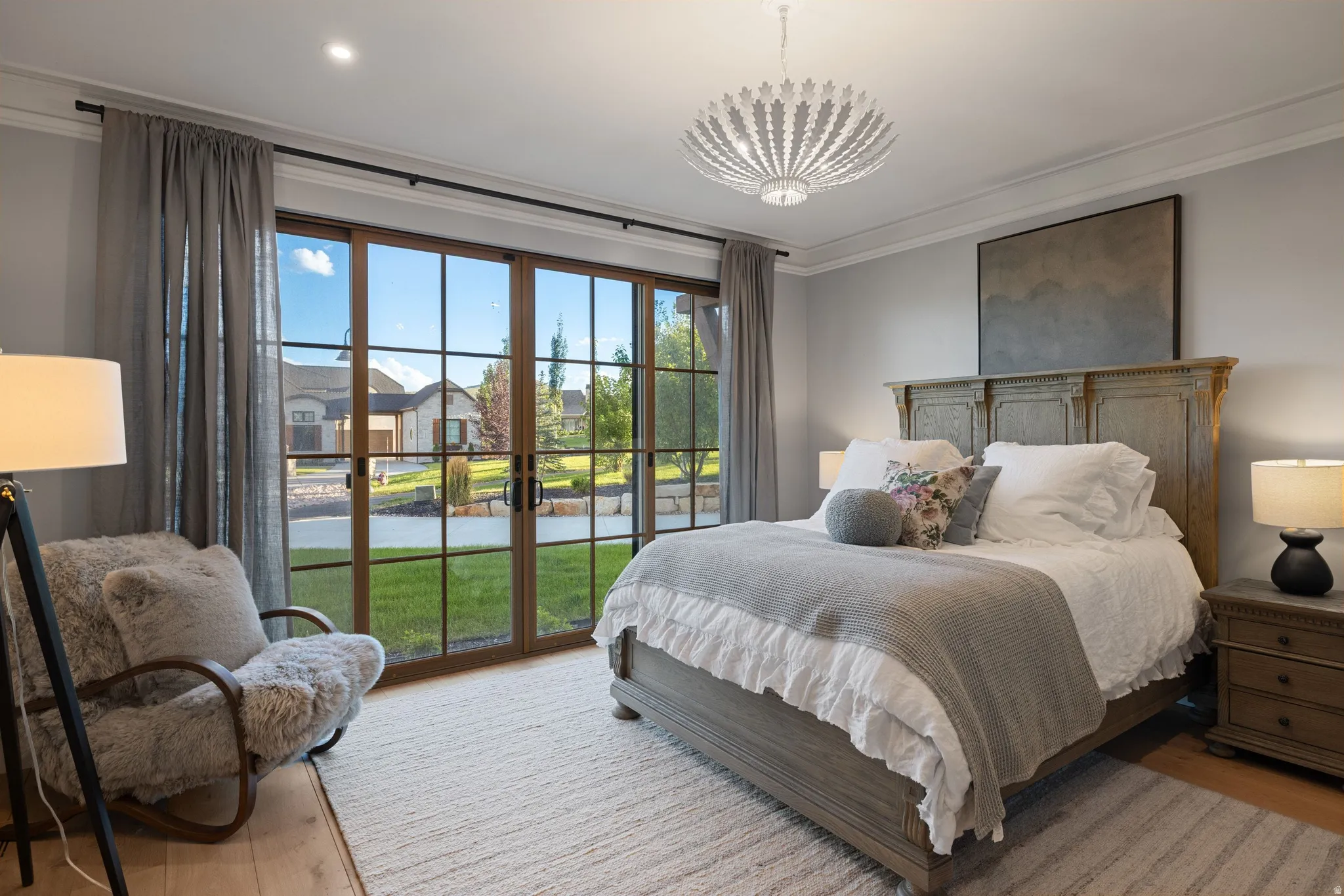 Primary bedroom featuring floor-to-ceiling glass doors opening to outdoor grounds, hardwood flooring, a wood-frame bed with upholstered headboard, dual nightstands with task lighting, decorative ceiling fixture, and natural light throughout the space