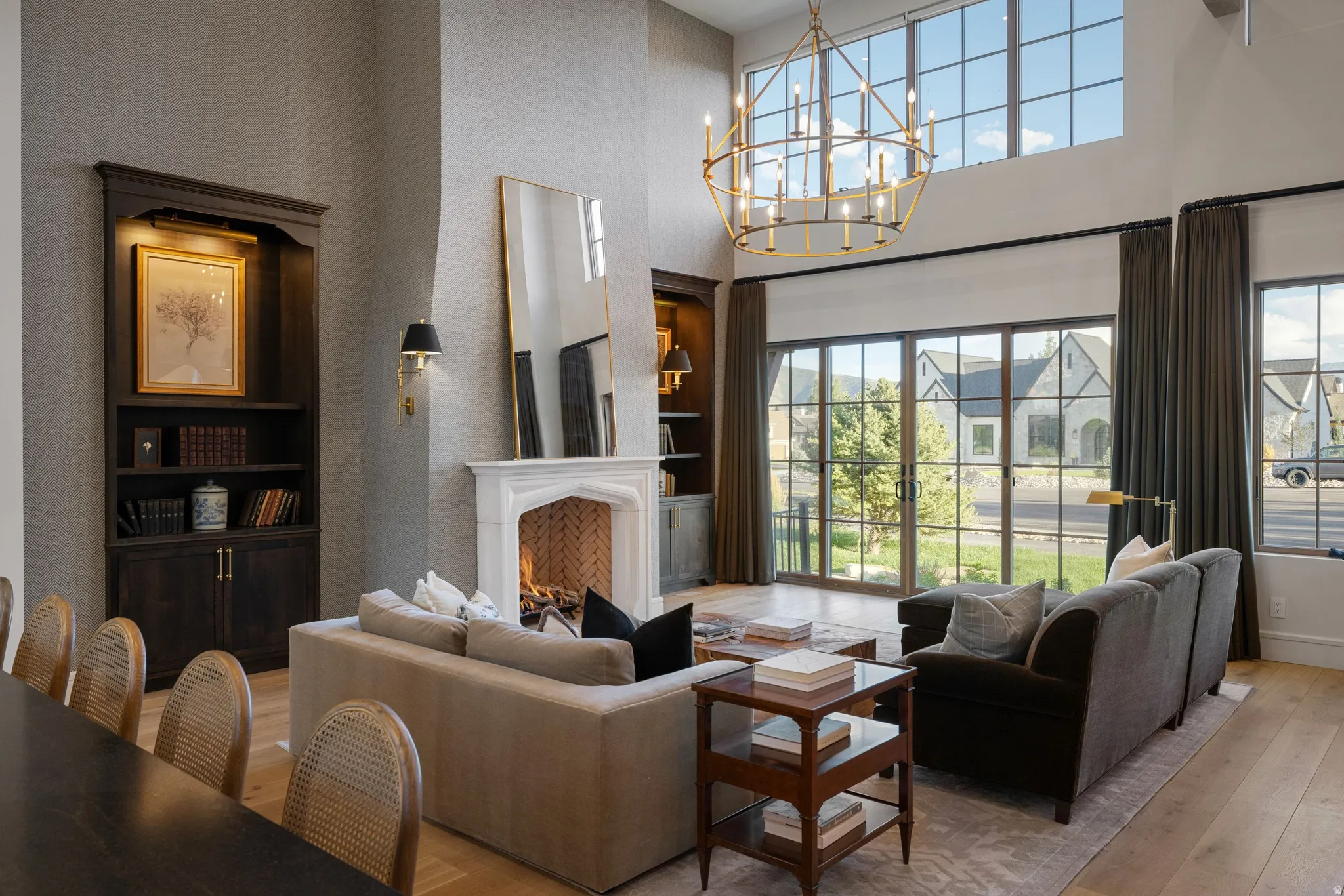 Sophisticated living space featuring soaring ceilings with an architectural brass pendant fixture, a wood-burning fireplace with white surround and dark frame accent, and expansive multi-pane glass doors opening to landscaped grounds