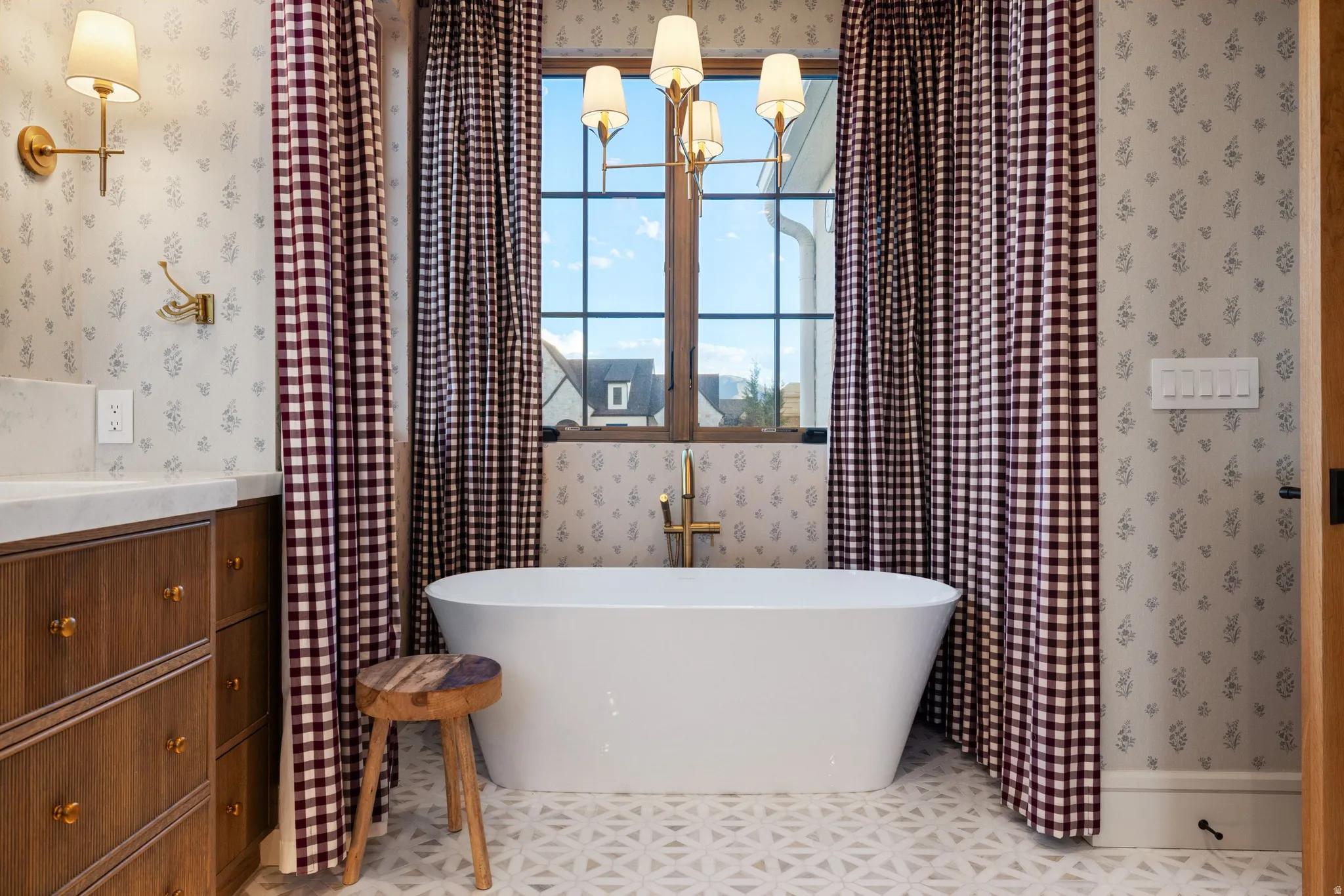 Freestanding soaking tub with floor-mounted brass faucet, patterned floor tiling, and a wooden vanity with a light-colored countertop