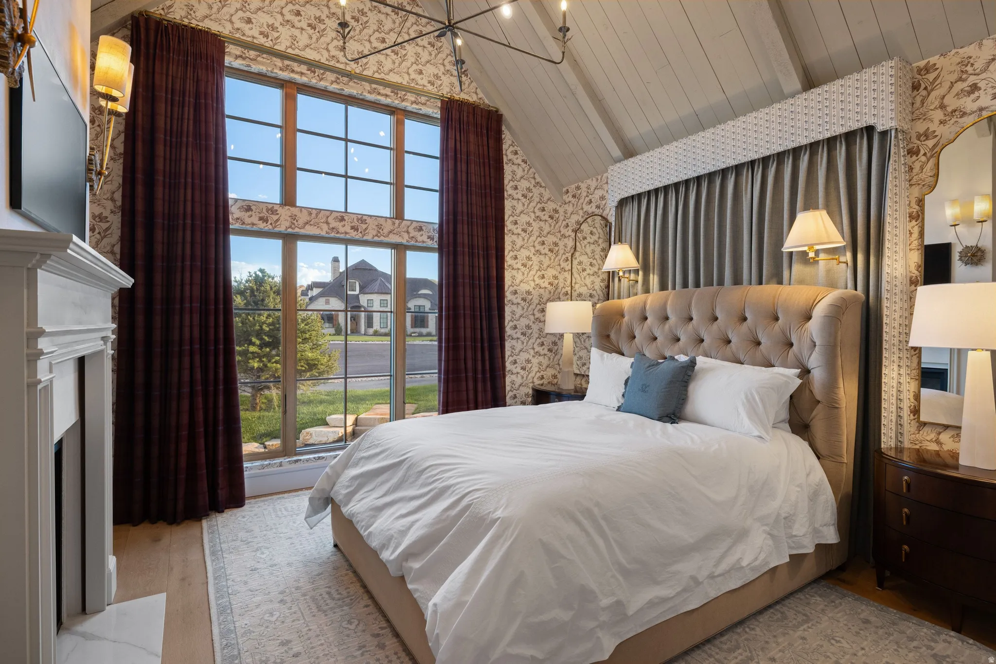Vaulted ceiling with exposed beams, wood-finish flooring, fireplace with white mantel, floor-to-ceiling windows, and patterned wallpaper