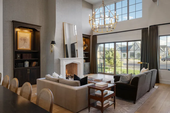 Sophisticated living space featuring soaring ceilings with an architectural brass pendant fixture, a wood-burning fireplace with white surround and dark frame accent, and expansive multi-pane glass doors opening to landscaped grounds