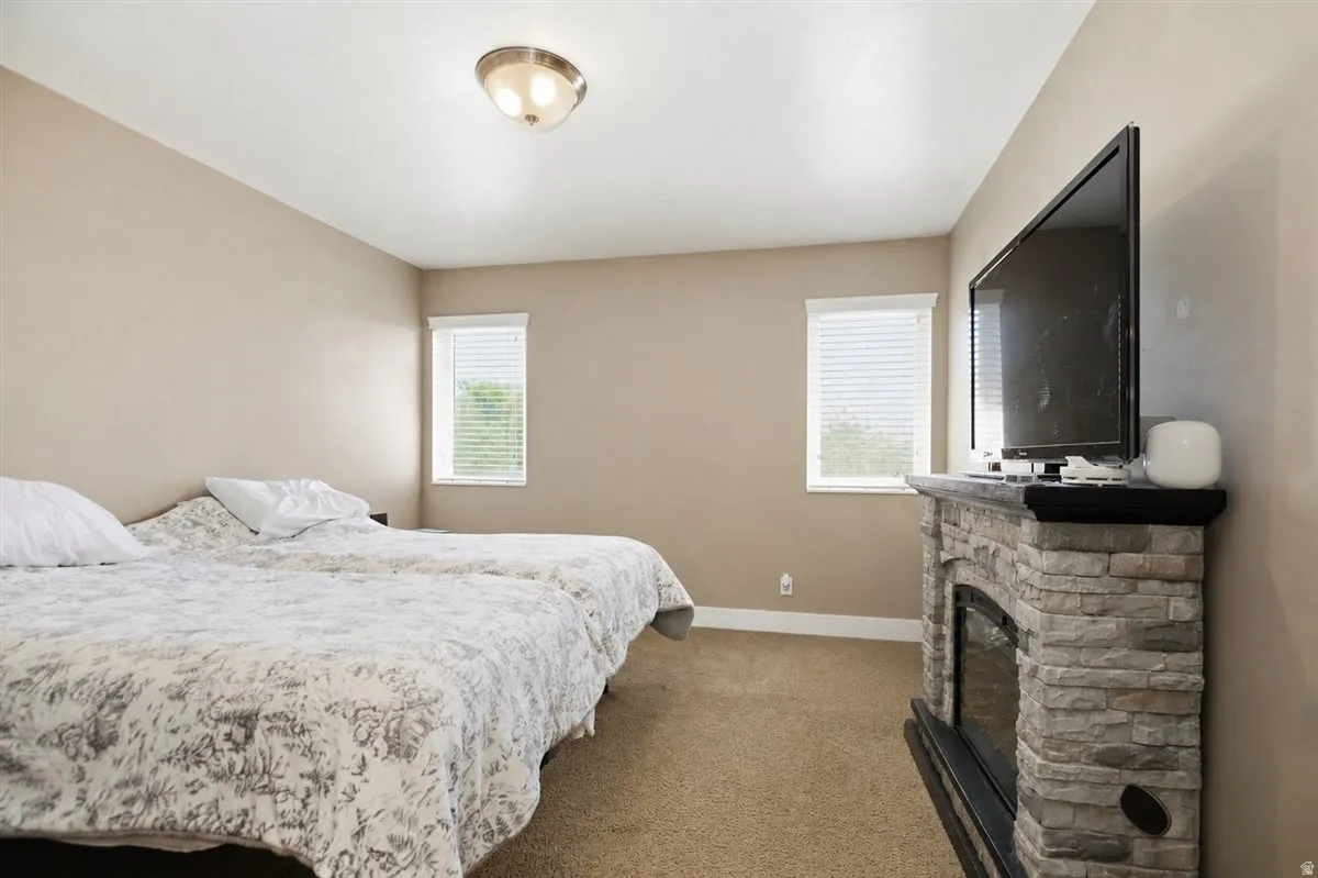 Carpeted flooring, two windows with blinds, a flush mount ceiling light, and a stone-clad fireplace with a dark mantel