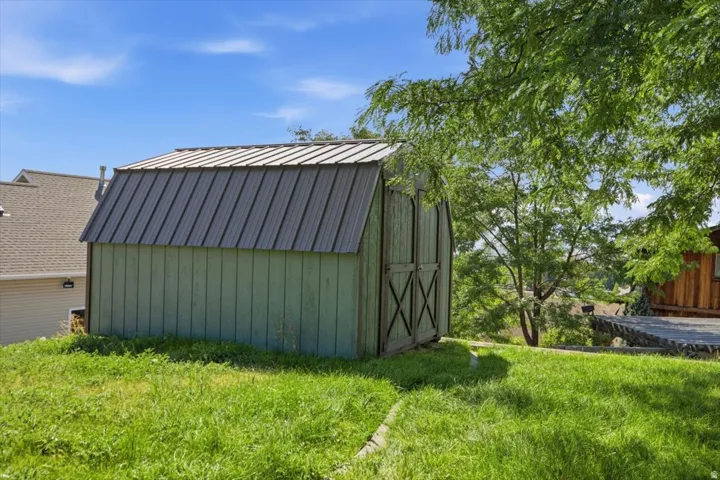 Green-sided utility shed with a metal roof and double doors, situated on a sloped grassy area