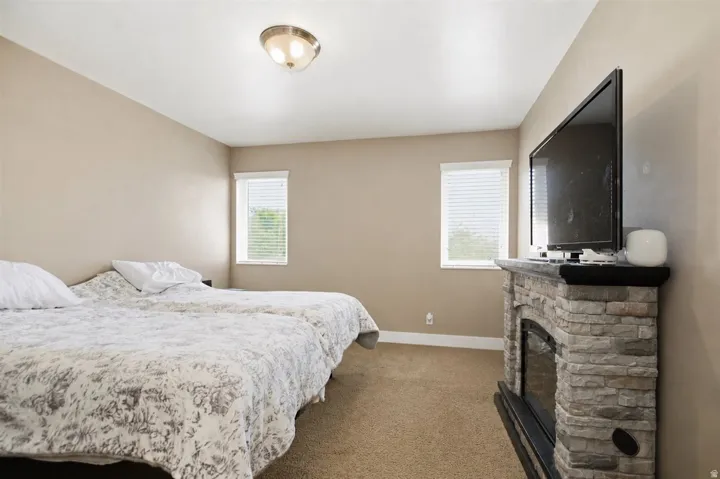Carpeted flooring, two windows with blinds, a flush mount ceiling light, and a stone-clad fireplace with a dark mantel